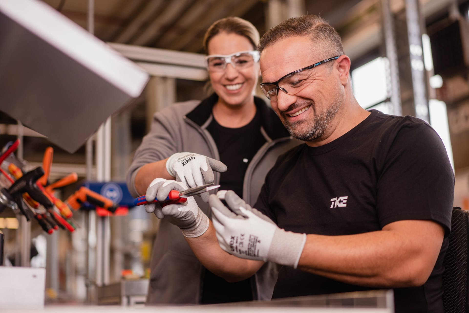 Eine Kollegin und ein Kollege mit Schutzbrillen und Arbeitshandschuhen begutachten lächelnd ein Werkzeug an der Werkbank.