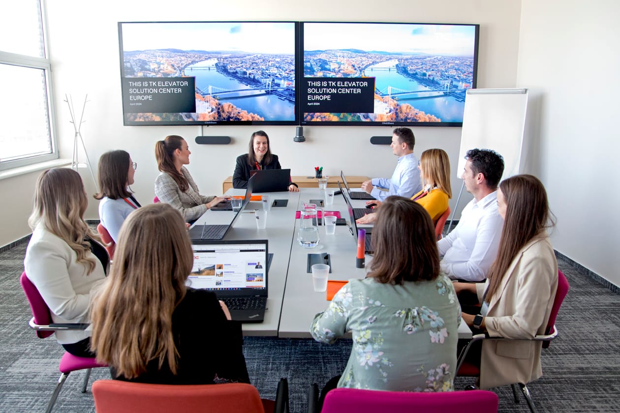 Ein Team bespricht sich an einem langen Konferenztisch unter zwei Bildschirmen mit dem Titel „This is TK Elevator Solution Center Europe“.