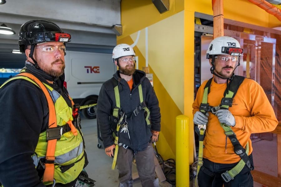 Drei TKE-Techniker mit Helm, Schutzbrille und Auffanggurt auf einer Baustelle.